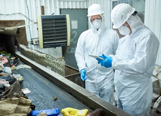 Portrait of two workers  wearing biohazard suits working at waste processing plant sorting trash and using digital tablet standing by conveyor belt, copy space